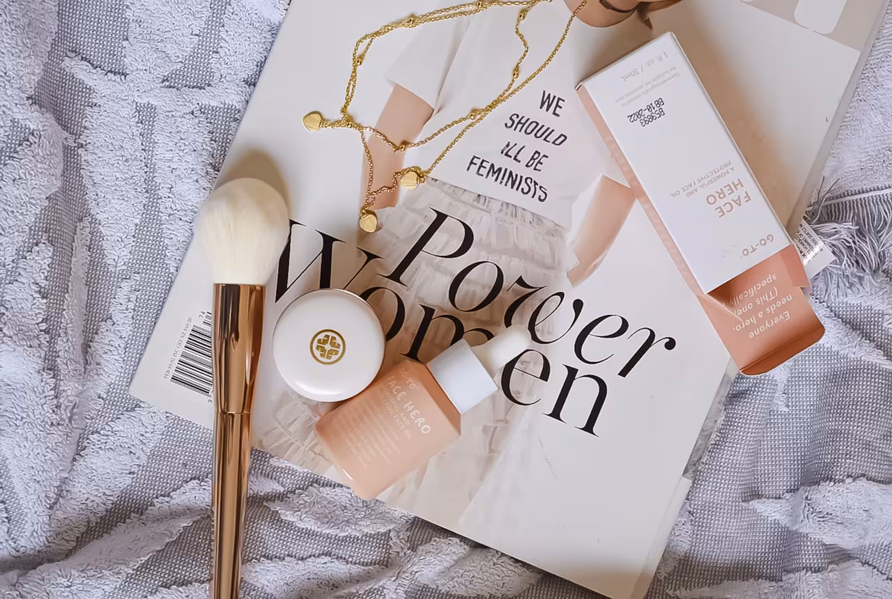 An open beauty PR box with crinkle paper and several unbranded skincare and makeup products arranged on a clean off-white surface, shot from above in soft daylight.