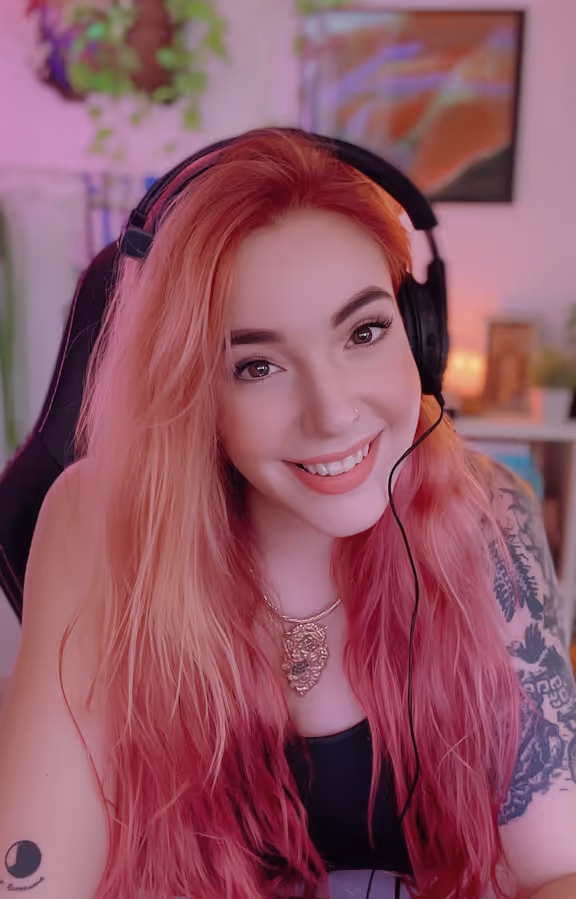 Smiling woman with long pink ombre hair wearing a headset and black top, seated in a gaming chair.